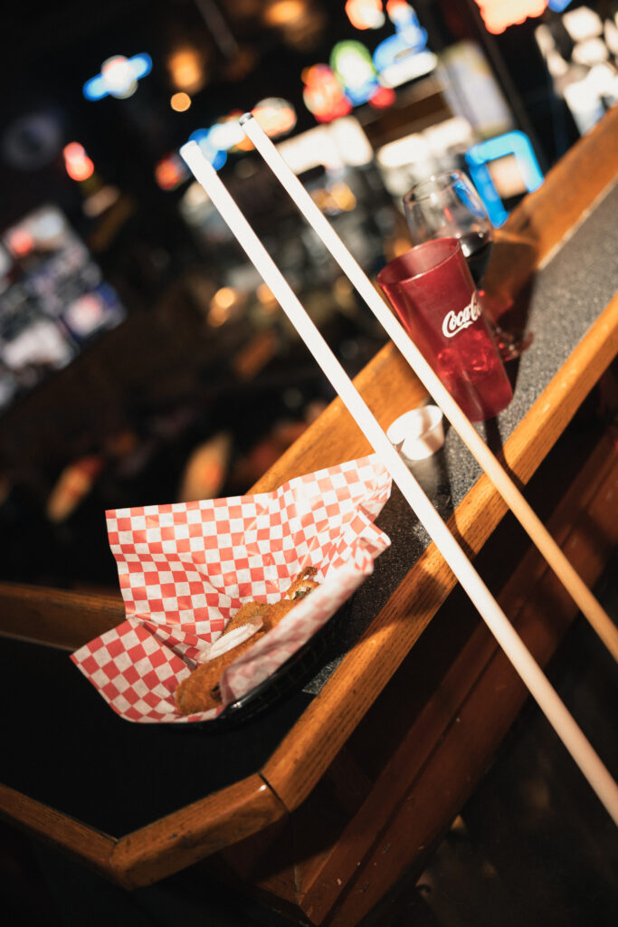 Crossed cue sticks resting beside checkered paper basket of fries on pool table rail