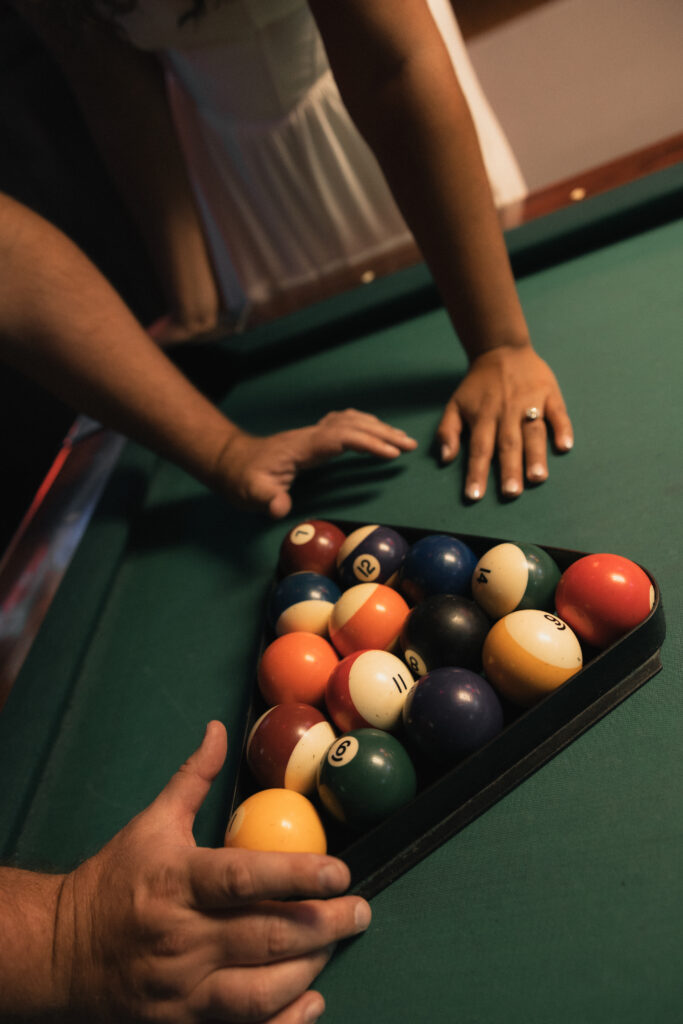 Close-up of hands over pool table, one hand flat with engagement ring visible, other racking balls into triangle