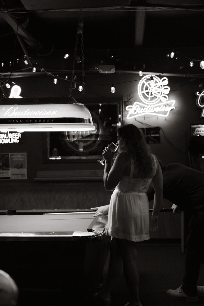 Black and white wide shot of Samantha standing at bar under glowing neon signage, Dave in background