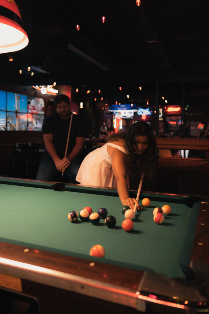 Wide shot of couple at pool table, Dave standing with cue stick, string lights and dartboard in background