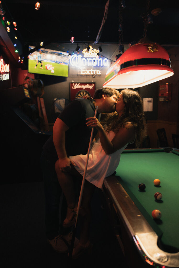 Samantha leaning over pool table to take a shot, Dave watching, neon Corona Extra sign overhead