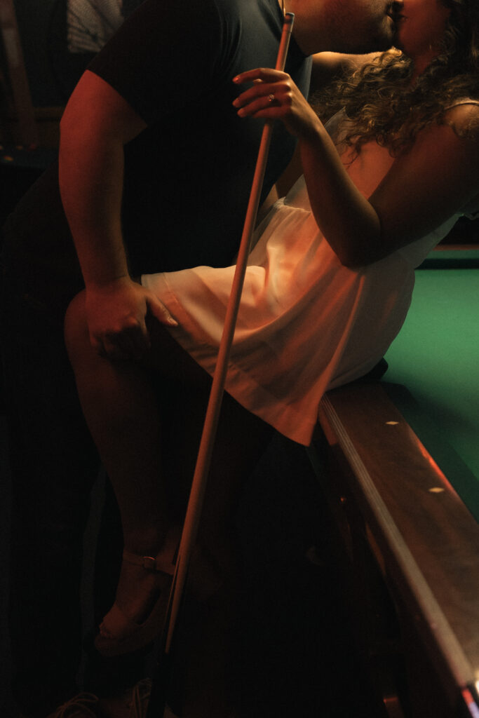 Samantha sitting on edge of pool table in white dress, warm ambient bar lighting