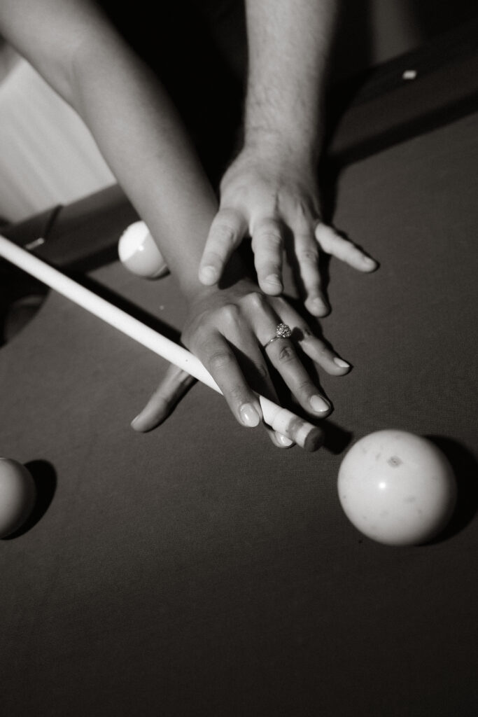 Black and white close-up of two hands gripping cue stick, engagement ring visible