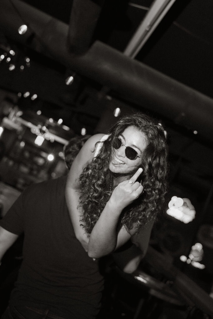 Black and white portrait of Samantha in sunglasses, curly hair loose, playful expression toward camera