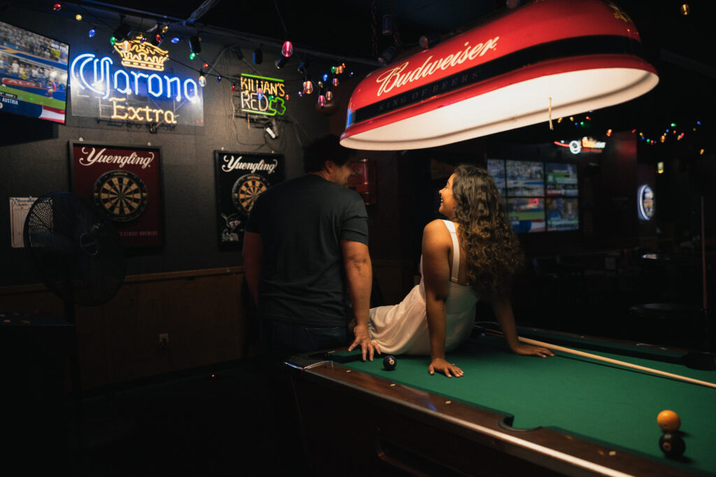 Dave leaning over pool table racking balls while Samantha leans against table in white dress, neon Budweiser sign glowing above