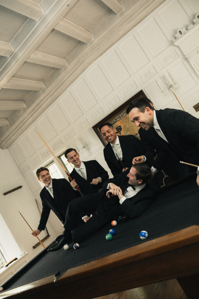Groomsmen laughing around pool table before wedding, NC estate venue
