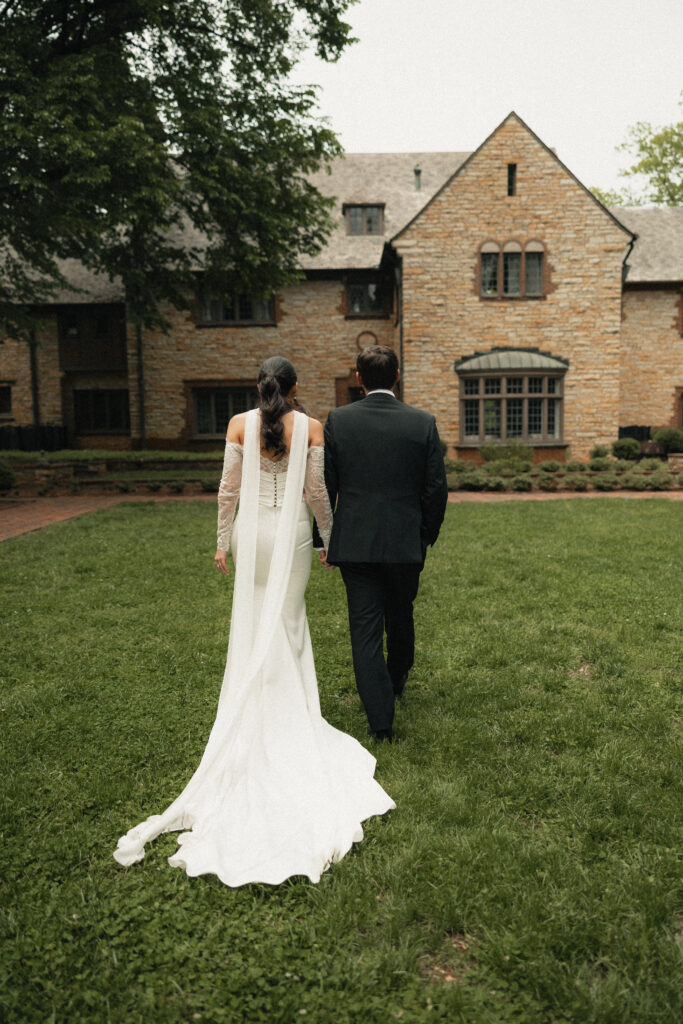 Bride and groom walking hand in hand across lawn, stone manor wedding venue