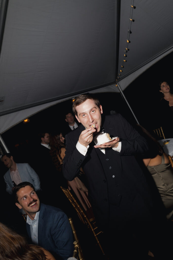 Groom eating cake slice under reception tent string lights