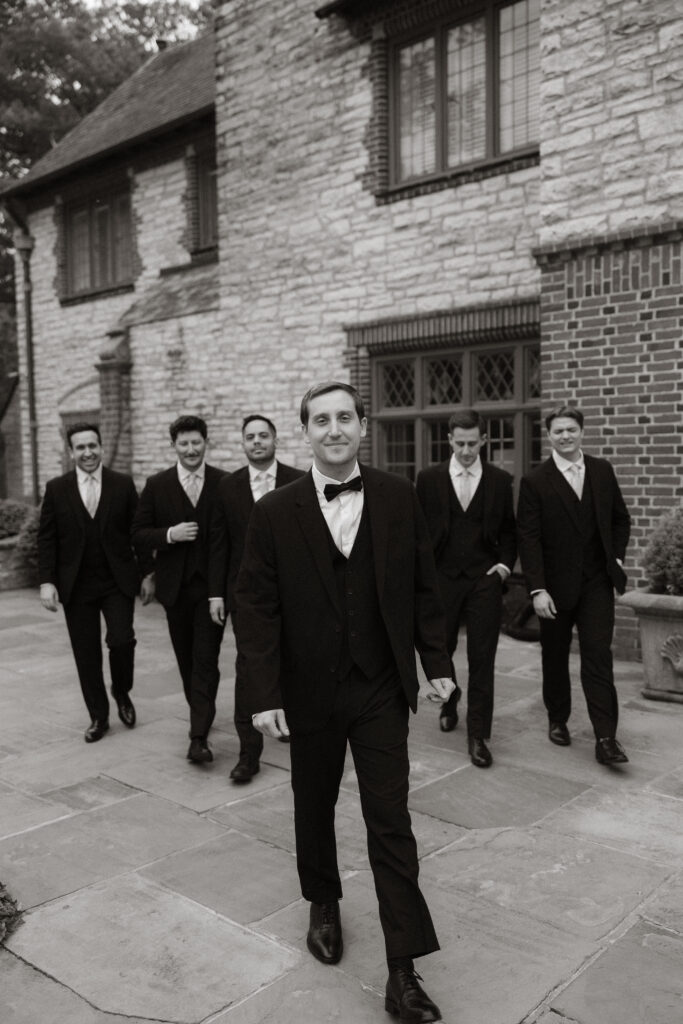 Groom and groomsmen walking outside stone manor, black and white portrait
